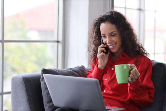 A woman looks at a laptop while talking on the phone and holding a coffee mug in her other hand