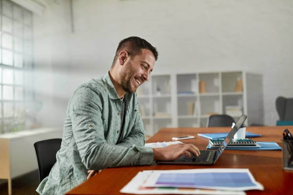 A man sits down smiling at a laptop as he types on it